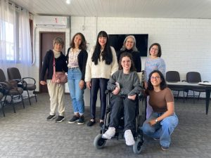 Fotografia colorida. Sete mulheres representantes da UFSC no V Fórum do Programa de Educação Profissional posam para foto sorrindo, distribuídas em duas fileiras. Atrás, em pé, da esquerda para a direita estão: Kamila (egressa da UFSC); Jéssica (servidora AI); Hilda (servidora AI); Karina (professora do curso de Administração); e Bianca (servidora CAE). Na frente estão Suélen (servidora AI) em sua cadeira de rodas e Tamara (servidora AI) agachada ao lado dela. Ao fundo se vê o saguão do auditório da FCEE com paredes e teto brancos, o piso cinza e algumas cadeiras encostadas nas paredes.