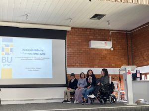 Fotografia colorida do palco do auditório da FCEE. À esquerda da imagem está a projeção da capa da apresentação de slides do AI, com a logo da biblioteca e informações sobre o AI e o evento. À direita estão Kamila; Bianca; Jéssica; e Suélen sentadas lado a lado. Jéssica fala ao microfone com o corpo voltado para a frente do palco. Ao fundo aparece a parede de tijolinhos e ao lado de Suélen um púlpito branco com o símbolo da FCEE em vermelho.