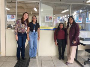 Fotografia colorida onde aparecem as servidoras da UFOPA e a servidora do AI, em pé e sorrindo para a câmera, em frente à entrada do setor. Da esquerda para a direita estão Juliane (UFOPA); Tamara (AI/BU); Ládia (UFOPA) e Monique (UFOPA). Atrás das mulheres está a divisória que delimita o setor, com a porta aberta.