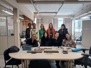 Fotografia colorida onde aparecem as servidoras e a estagiárie do NDI, juntamente com as servidoras e estagiárias do AI, dentro da sala do setor. Em pé, sorrindo para a câmera, da esquerda para a direita, estão: Maria António (AI), Shirlei (AI), Vandiara (AI), Lídia (AI) e Hilda (AI). Sentadas à frente, também sorrindo para a câmera, da esquerda para a direita, estão: Niteri (NDI), Luz (NDI), Katia (NDI) e Suélen (AI). À frente delas há uma mesa de reunião oval, com alguns equipamentos de tecnologias assistivas expostos. Ao fundo, é possível ver computadores, janelas com cortinas e a porta do setor. Fim da descrição.