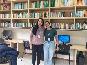 Fotografia colorida onde aparecem as servidoras Hilda e Tamara, em pé e sorrindo para a câmera, em frente à uma estante aérea cheia de livros. Abaixo da estante há uma mesa comprida com cadeiras pretas e computadores. Fim da descrição.